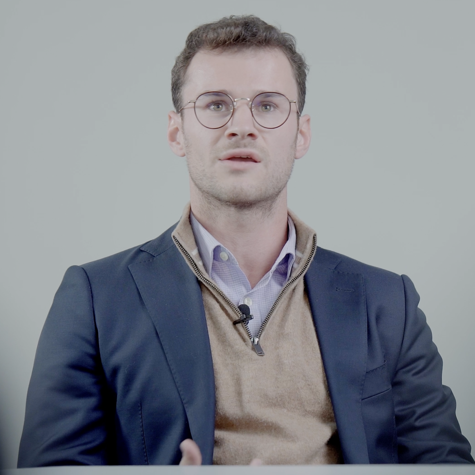 A person in a blazer and sweater sits at a table, hands gesturing, with a neutral background.