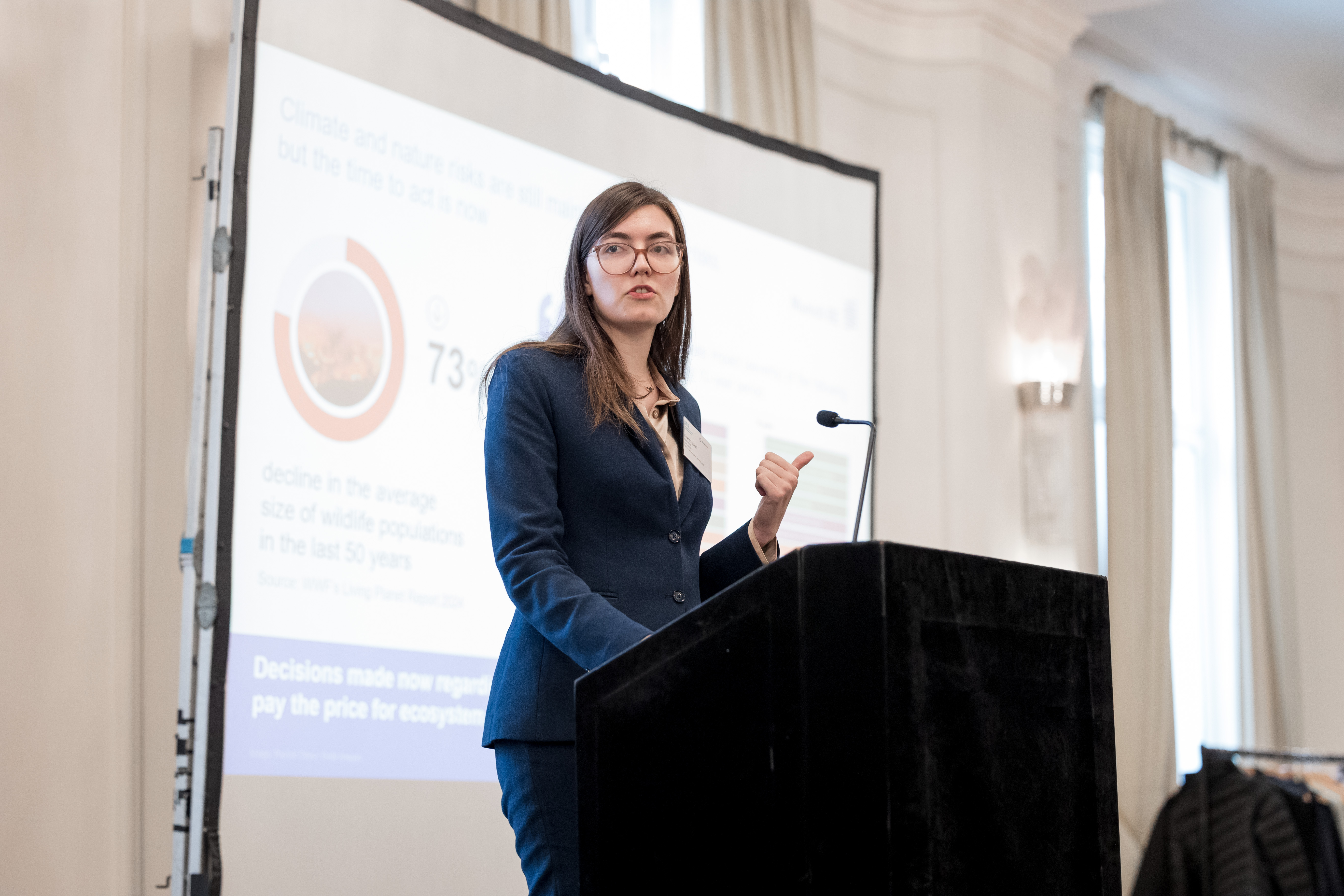 A woman in a suit speaks at a podium, with a presentation slide visible behind her.