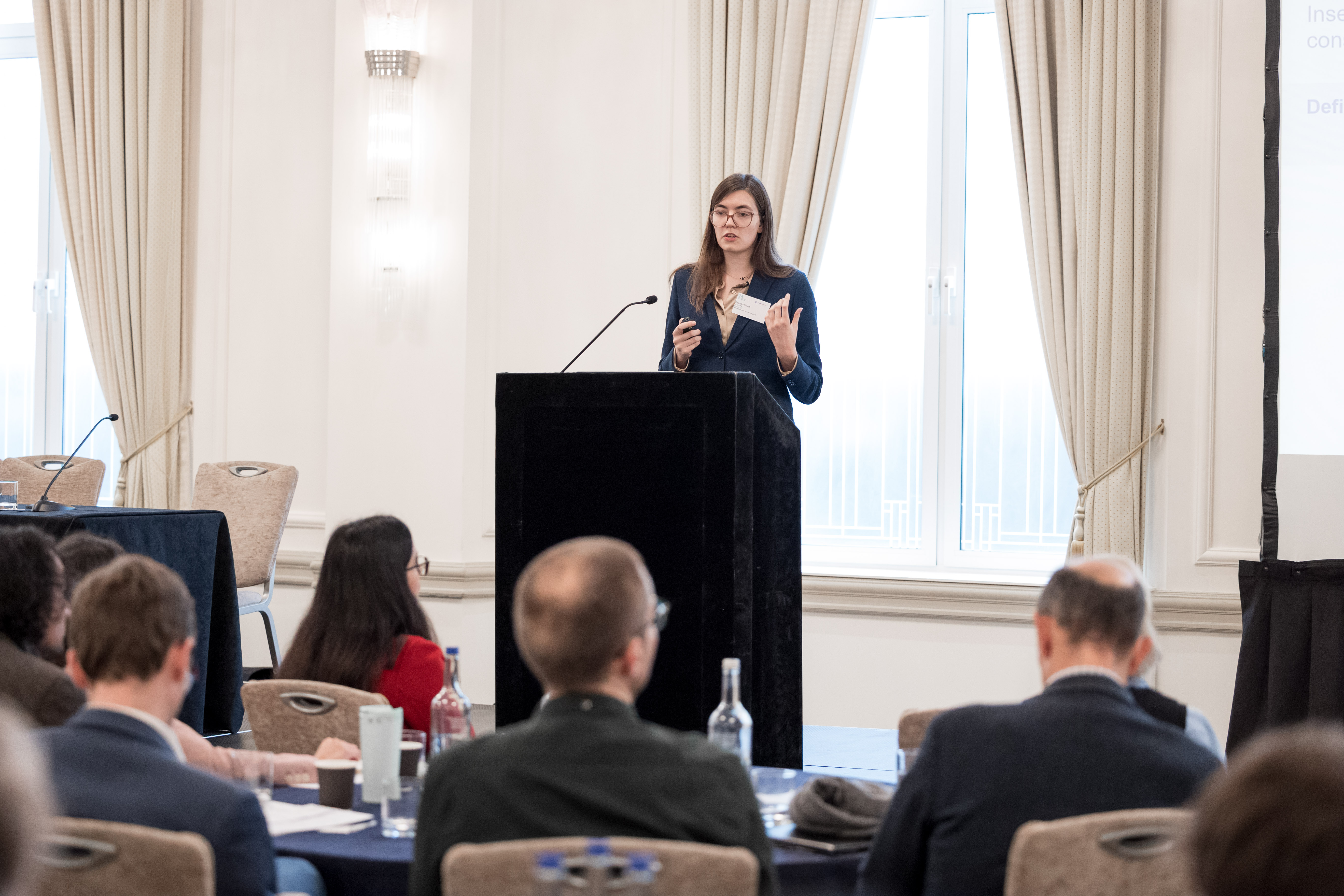 A speaker presents at a podium in a conference room, with an audience seated at tables in the foreground.