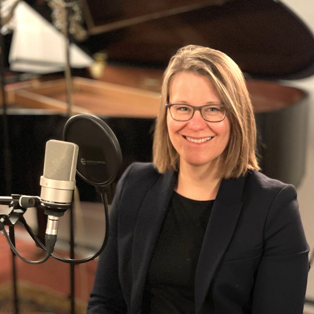 A person sits in front of a microphone in a recording studio with a grand piano in the background.