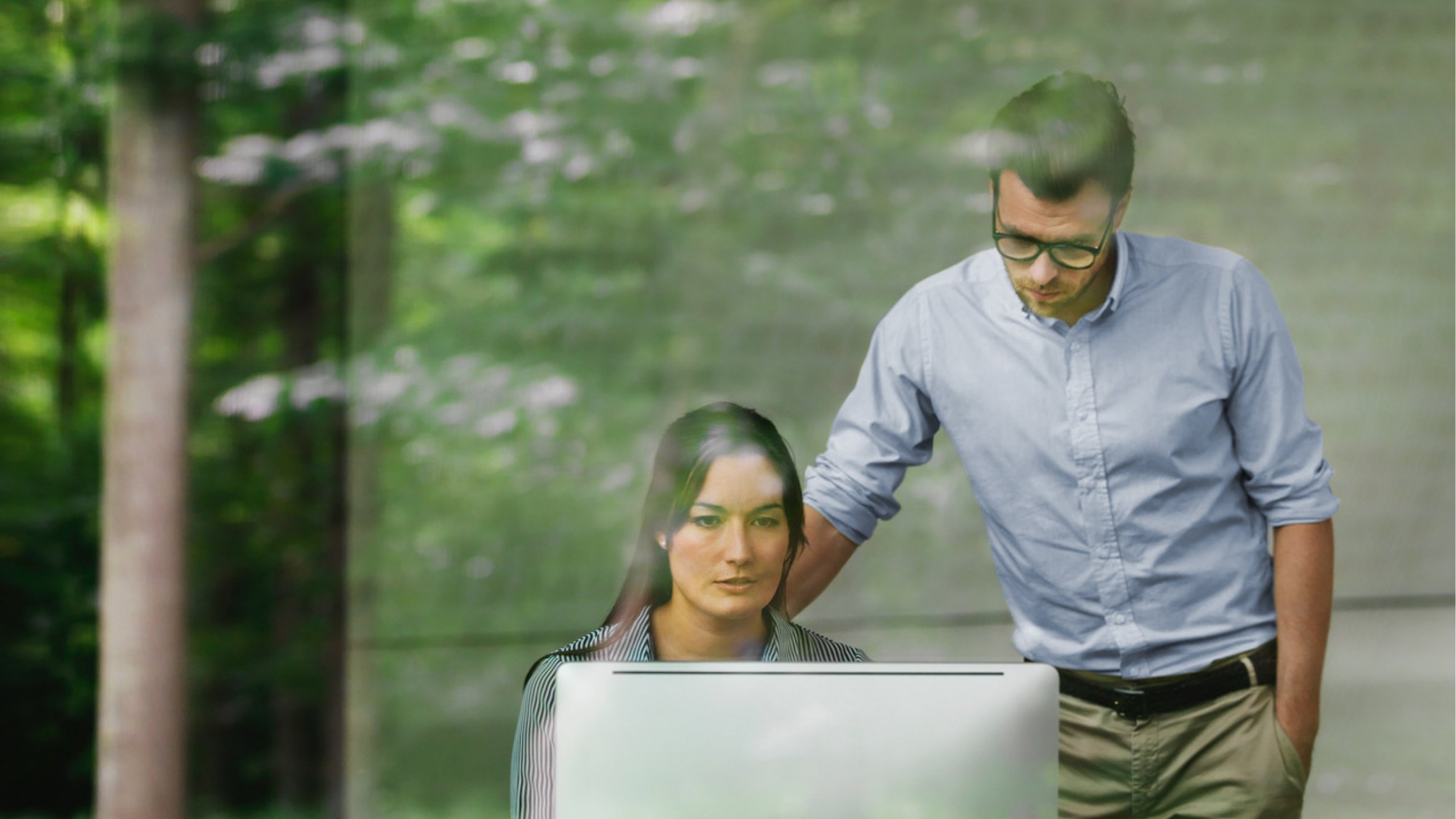 A man stands beside a woman at a computer, with a green forest visible through a large window behind them.