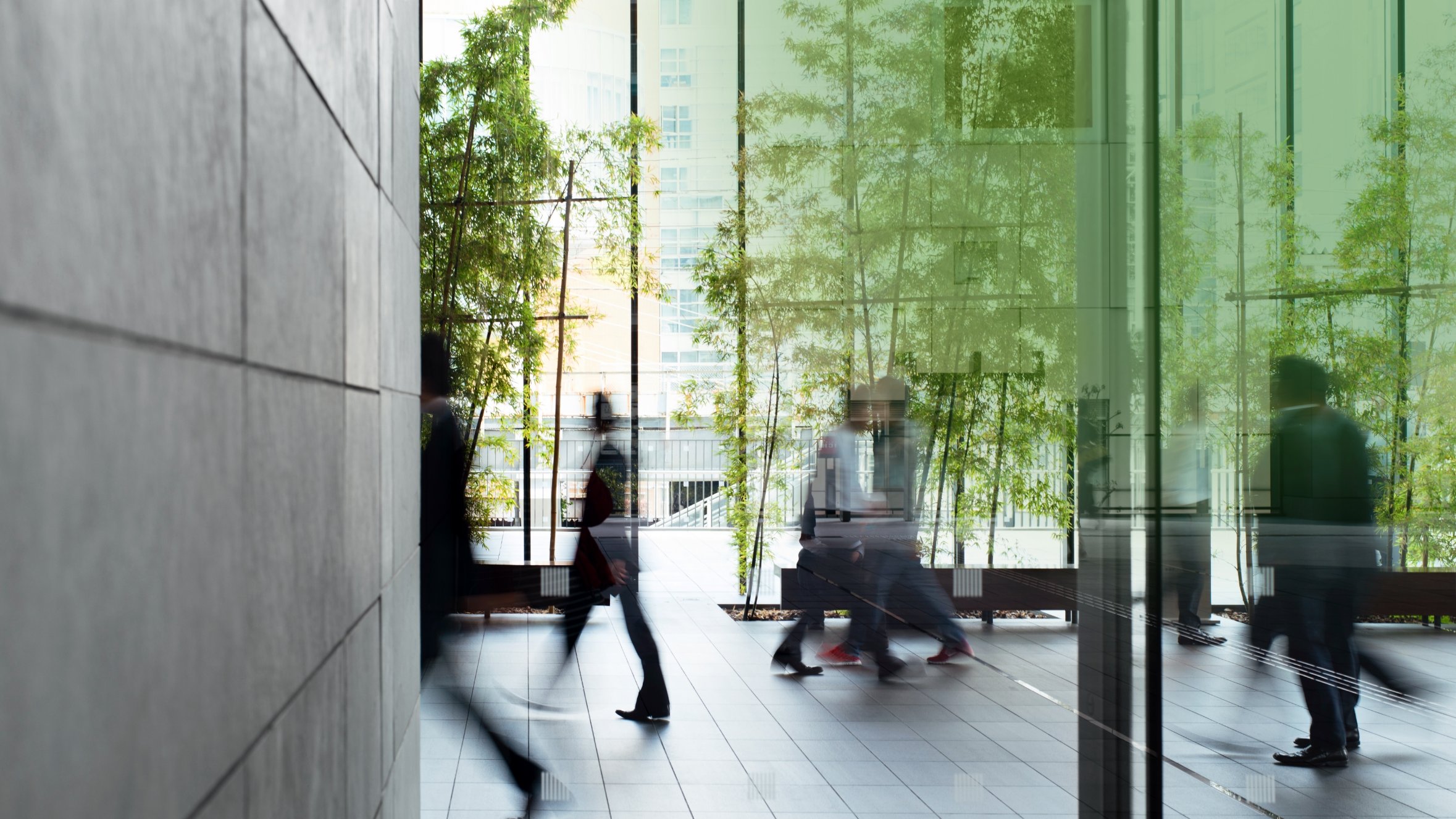 People walking in a modern building with glass walls and greenery visible in the background.