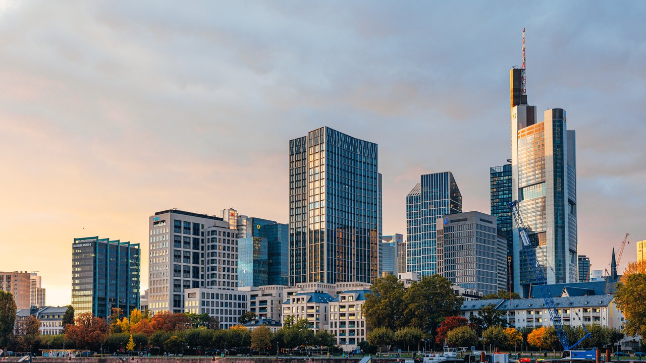 A skyline view of modern buildings by a river during sunset, with trees and a construction crane in the foreground.