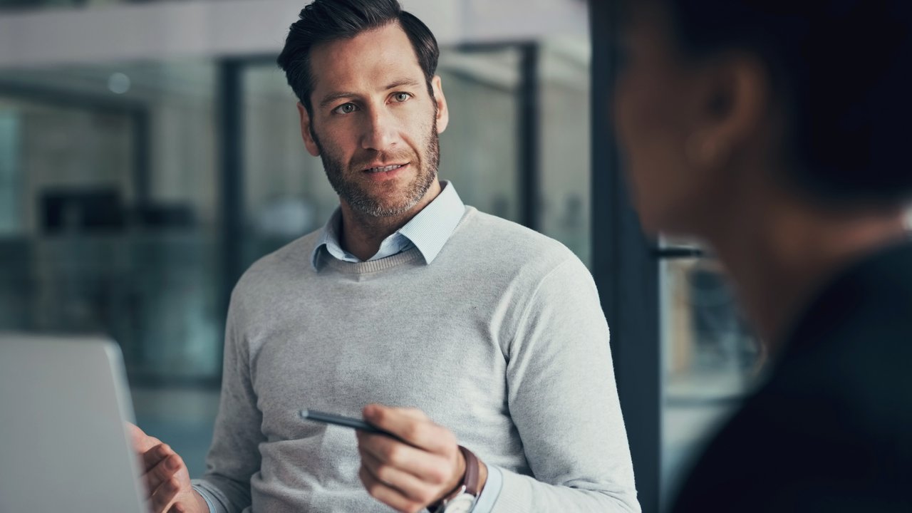 A man in a gray sweater speaks while holding a pen, seated at a table with a laptop.
