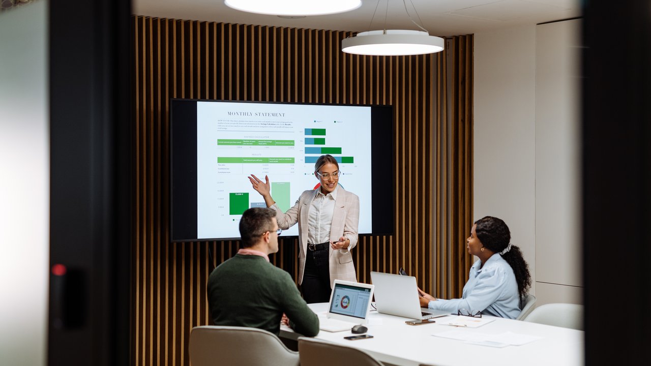 A business meeting with three people discussing a presentation on a screen displaying graphs and data.
