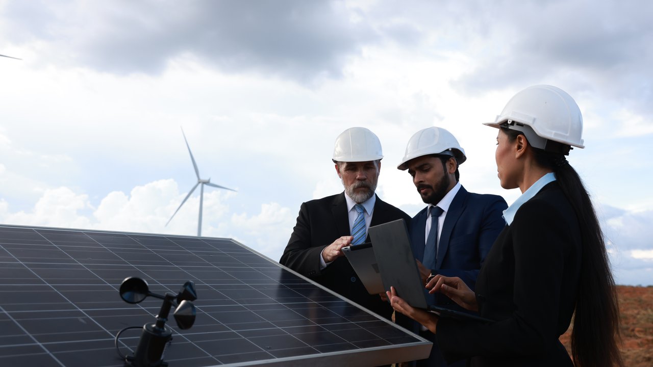 Three professionals in hard hats examine a solar panel and a laptop, with wind turbines in the background.
