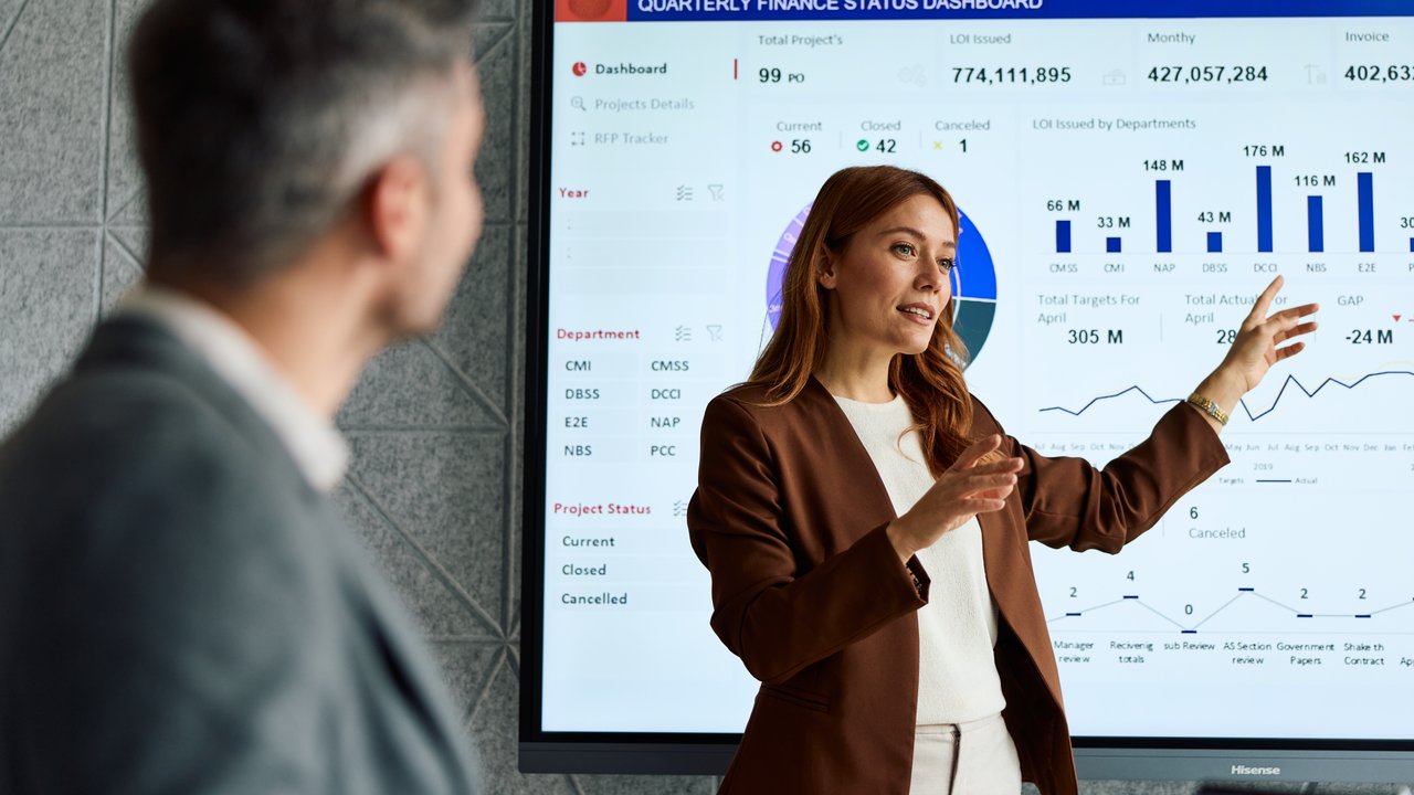 A woman presents data on a finance dashboard while a man listens. The screen displays charts and project statuses.
