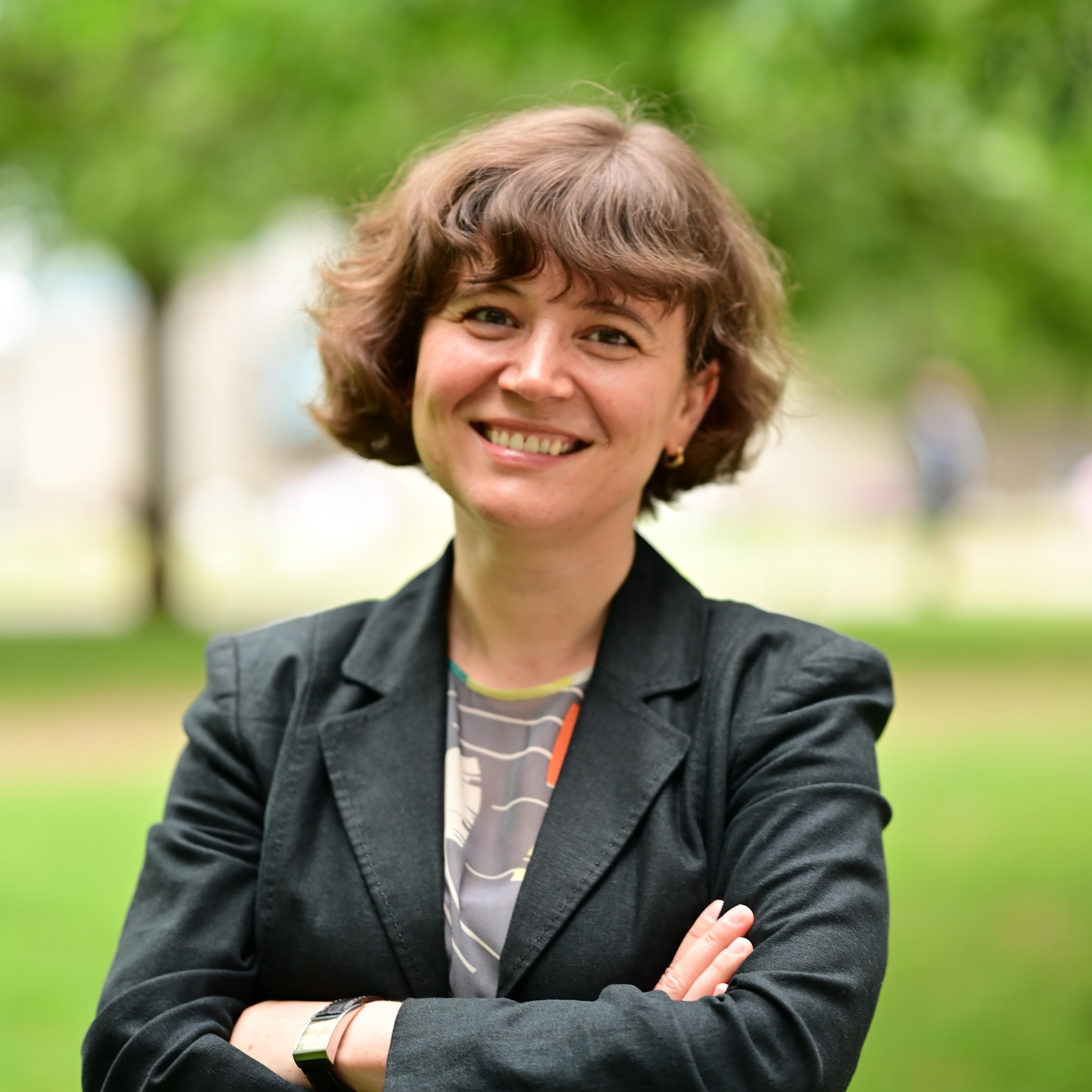 A person with short, curly hair stands outdoors, wearing a black blazer and a patterned shirt, arms crossed.