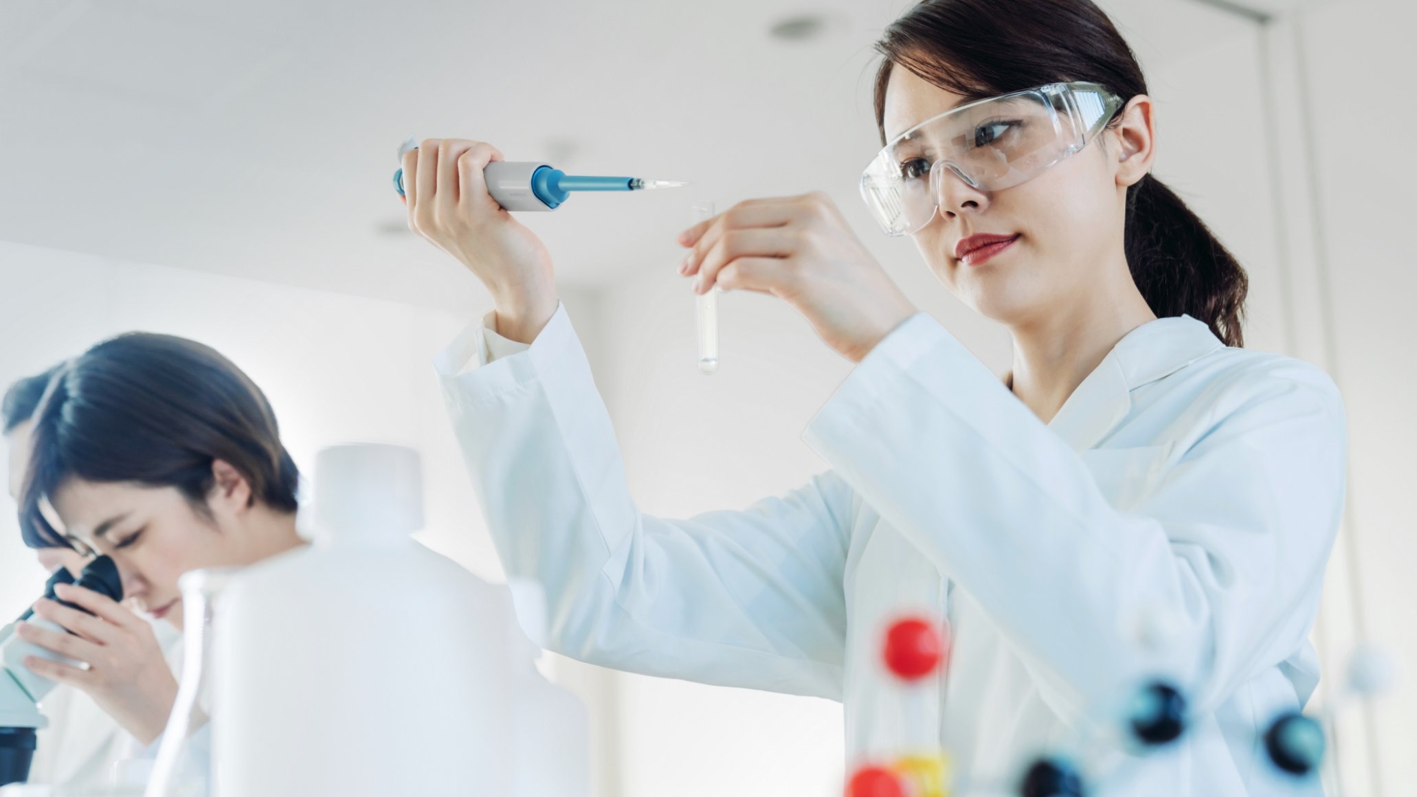 Two scientists in lab coats work with test tubes and a microscope in a bright laboratory setting.
