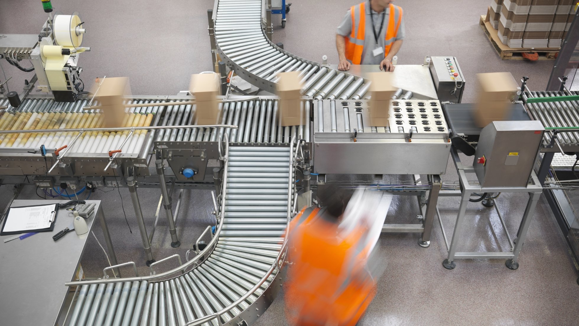 A conveyor system in a warehouse with workers in orange vests handling boxes and equipment.